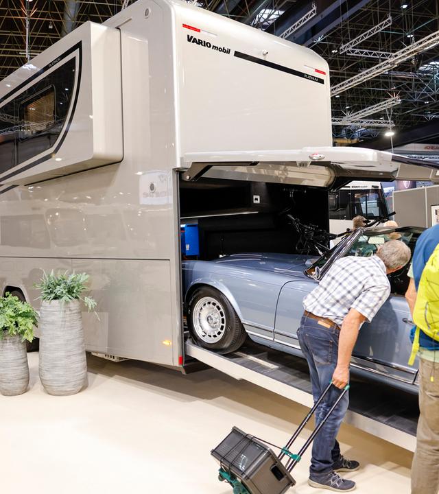 Zwei Messebesucher betrachten ein Wohnmobil, in dessen Heck ein Auto wie in eine Garage hineinfährt.
