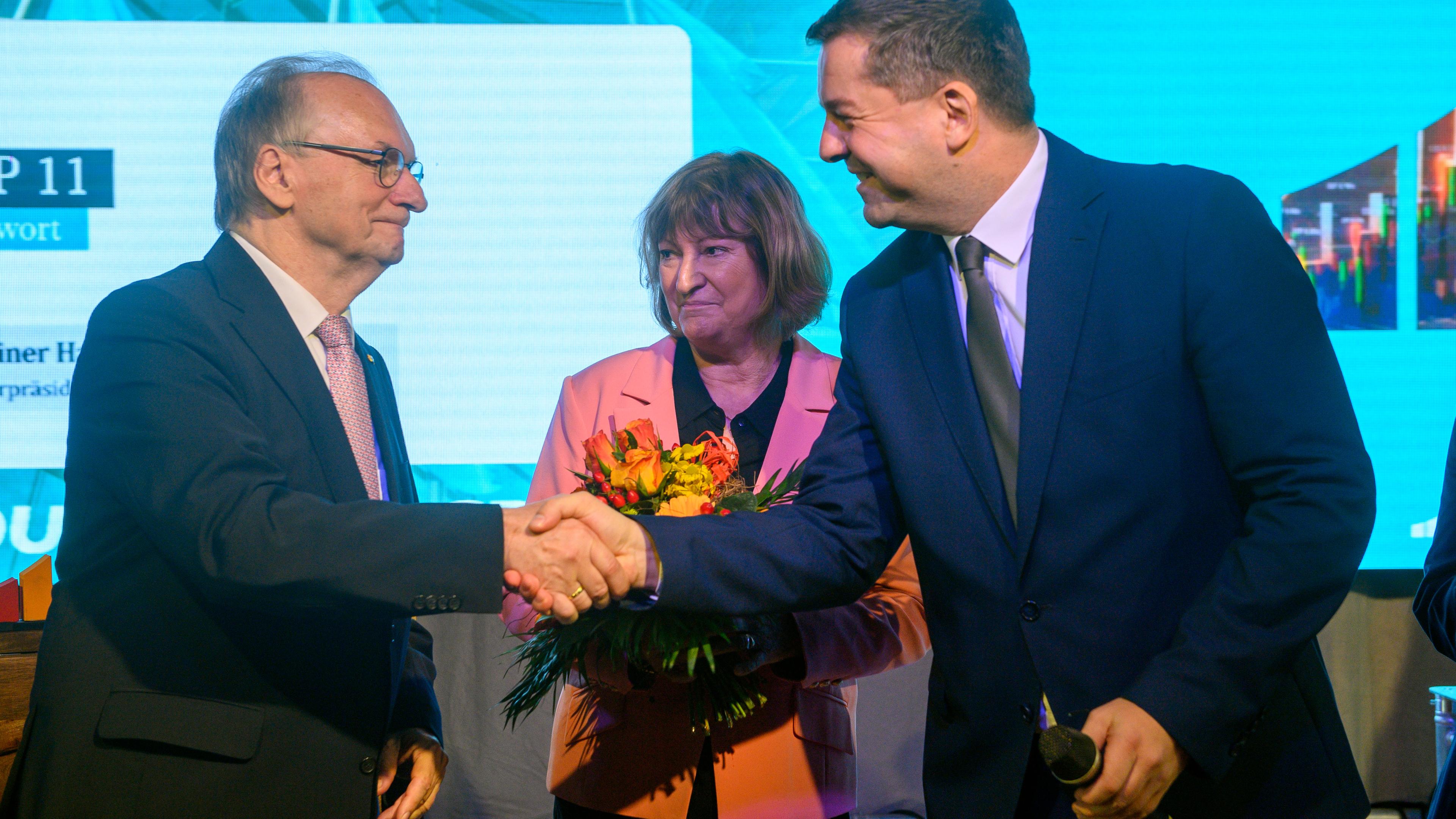 Reiner Haseloff (l-r, CDU), Ministerpräsident des Landes Sachsen-Anhalt und Ehefrau Gabriele Haseloff sowie Sven Schulze (CDU), Minister für Wirtschaft, Tourismus, Landwirtschaft und Forsten des Landes Sachsen-Anhalt stehen mit Blumen auf der Bühne der Landesvertreterversammlung der CDU und geben sich die Hände.