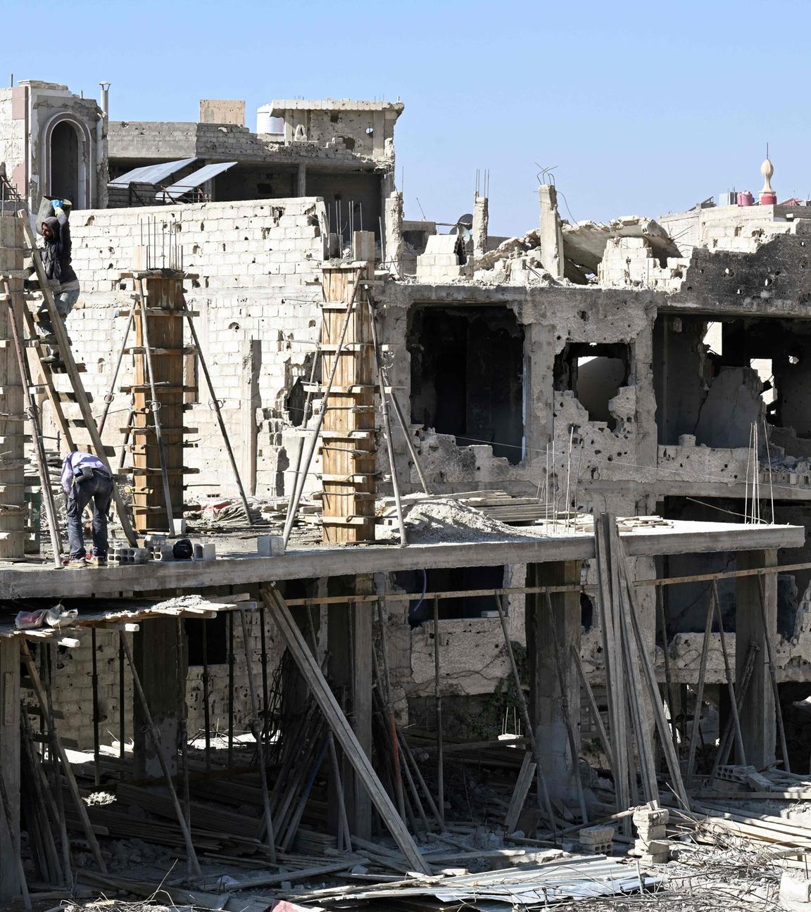 Ruinen von Häusern mit vereinzelten Gerüsten daran vor blauem Himmel.