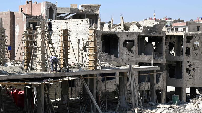 Ruinen von Häusern mit vereinzelten Gerüsten daran vor blauem Himmel.