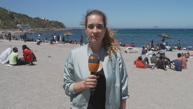 ZDF-Reporterin Scarlett Sternberg vom Strand bei Ceuta. Hinter ihr sieht man viele männlich-aussehende Menschen, die größtenteils in kleinen Gruppen rumsitzen.