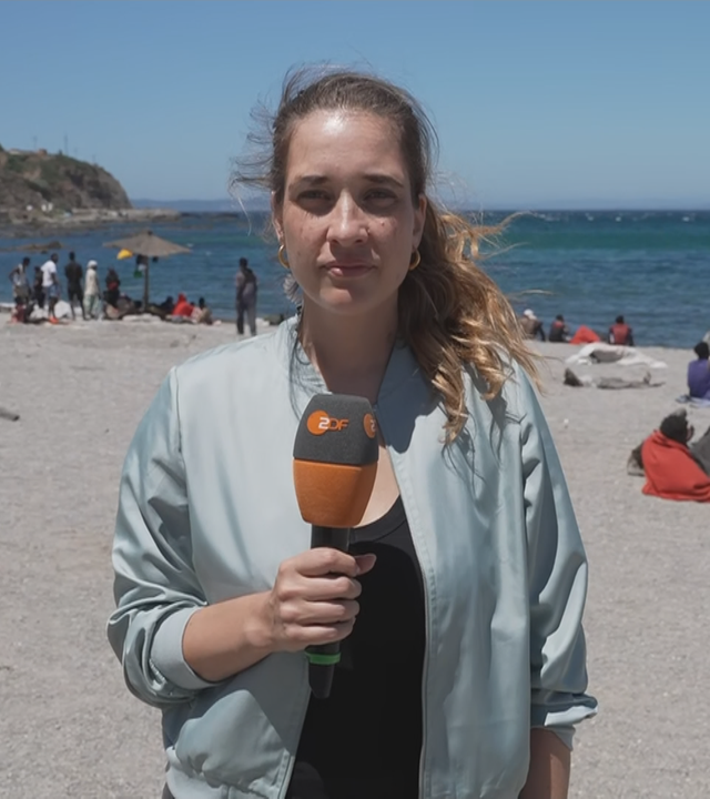 ZDF-Reporterin Scarlett Sternberg vom Strand bei Ceuta. Hinter ihr sieht man viele männlich-aussehende Menschen, die größtenteils in kleinen Gruppen rumsitzen.
