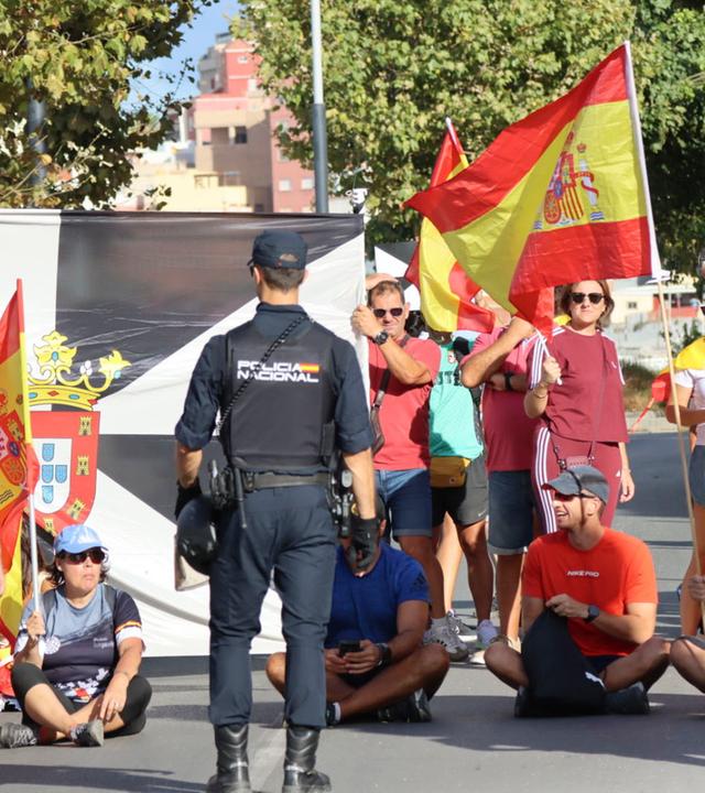 Einwohner von Ceuta sitzen in einer Reihe auf dem Boden, sie schwenken spanische Flaggen. Davor stehen Polizisten.