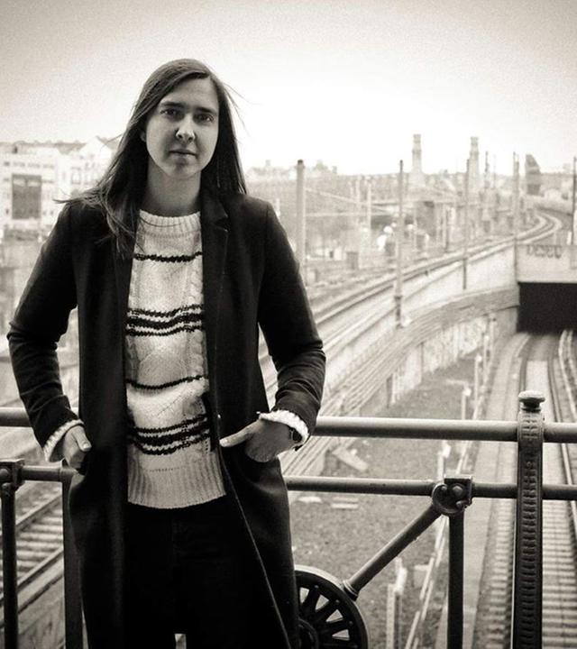 Die Saxophonistin Sarah Chaksad auf einer Bahnhofsbrücke, beide Hände seitlich in den Hosentaschen vom Körper, in die Kamera schauend. Hinter ihr die Gleise.