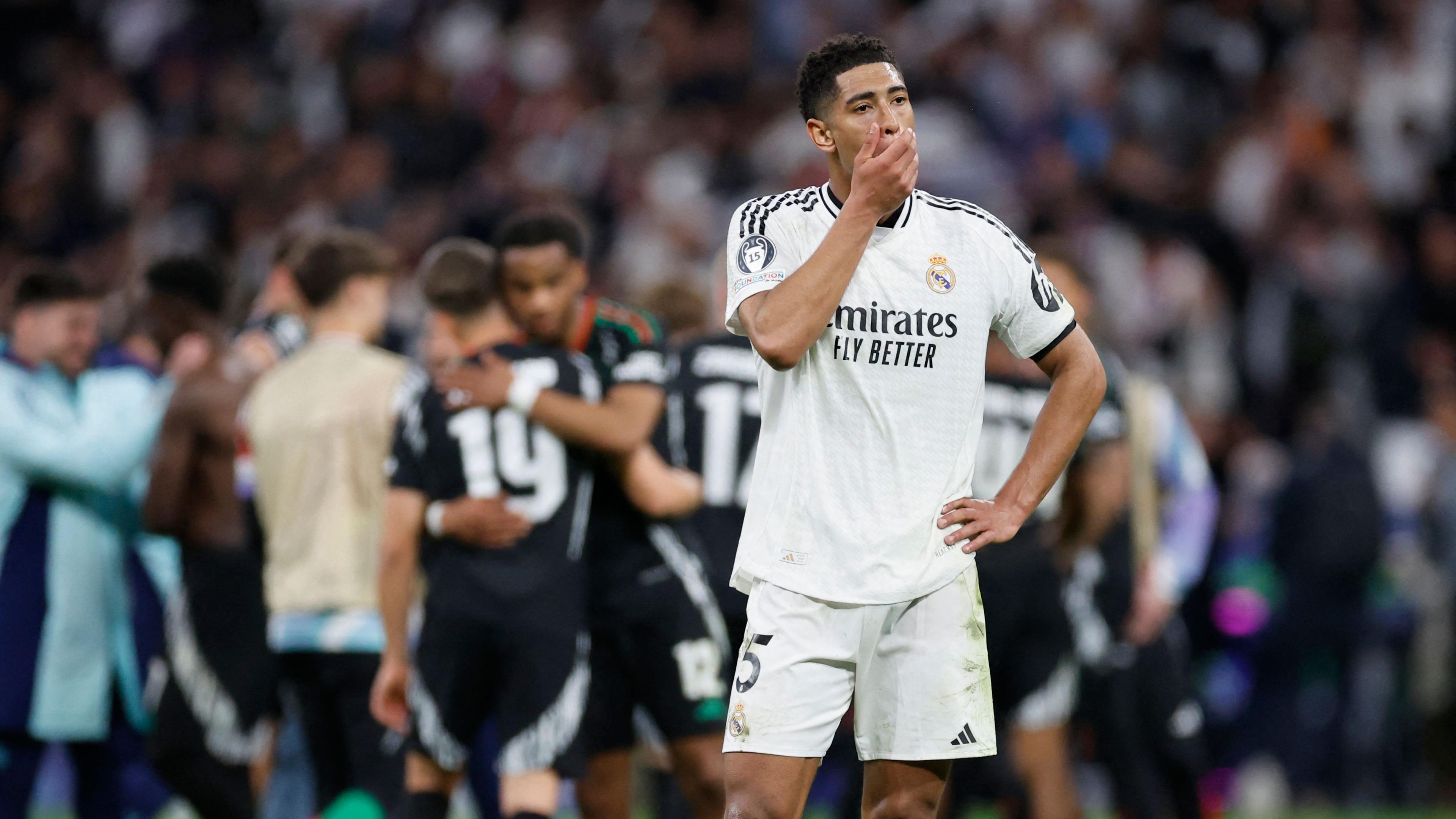 Real Madrids Jude Bellingham steht enttäuscht auf dem Rasen, während die Spieler von Arsenal FC im hintergrund jubeln.