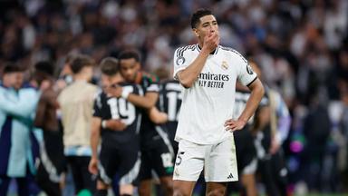 Real Madrids Jude Bellingham steht enttäuscht auf dem Rasen, während die Spieler von Arsenal FC im hintergrund jubeln.