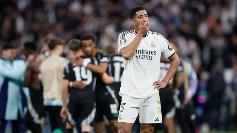 Real Madrids Jude Bellingham steht enttäuscht auf dem Rasen, während die Spieler von Arsenal FC im hintergrund jubeln.