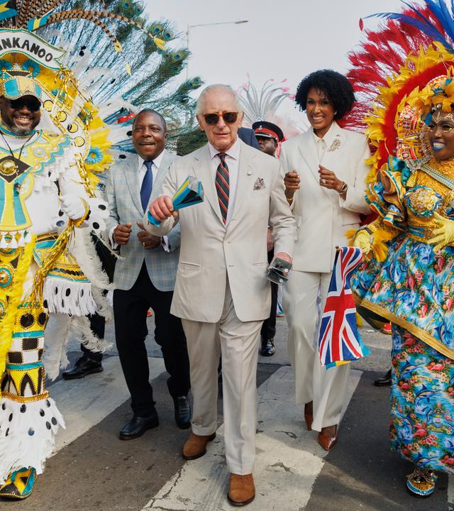 Der britische König Charles III.schüttelt traditionelle Kuhglocken, während er mit Landwirtschaftsminister Jomo Campbell (links) und Kulturministerin Leslia Brice (rechts) zwischen Junkanoo-Tänzern läuft. 