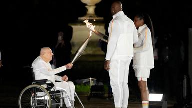 Charles Coste übergibt bei der Eröffnungsfeier der Olympischen Sommerspiele in Paris 2024 im Rollstuhl sitzend das olympische Feuer