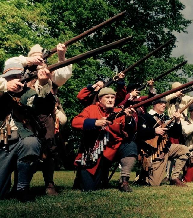 Auf einer Wiese kniet eine Gruppe von Männern in einer Reihe. Sie sind mit Musketen bewaffnet und halten die Gewehre im Anschlag.