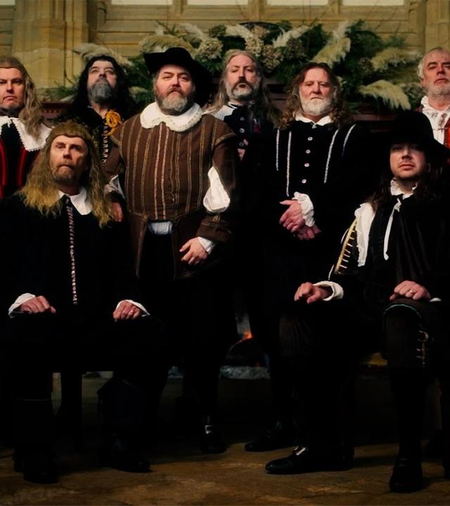 Spielszene: Sechs Männer in historischen Kostümen stehen vor einer holzgetäfelten Wand. Vor ihnen sitzen zwei weitere Männer. Alle tragen dunkle Wamse mit weißen Krägen und Manschetten sowie dunkle Hosen und schwarze Schuhe mit Schnallen.