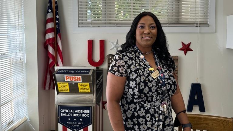 Eine Schwarze Frau steht im Kleid und mit vielen Anstecknadeln an einem Band vor einer Wahlurne in einem hellen Raum.