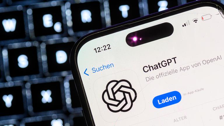 Ein Smartphone mit der App des KI-Chatbots ChatGPT ist vor dem Logo des US-Softwareunternehmens OpenAI zu sehen