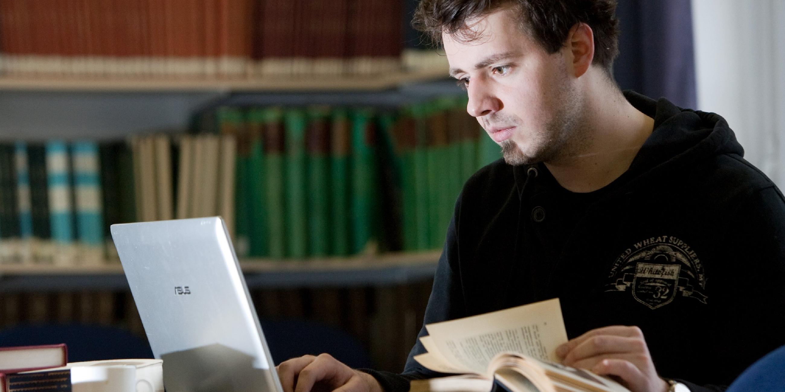 Student sitzt in einer Bibliothek und arbeitet mit Laptop und Büchern.