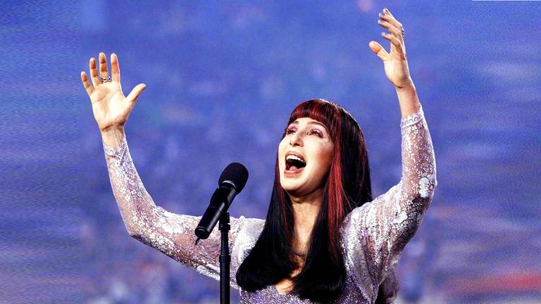 Cher steht auf der Bühne und singt mit erhobenen Armen 