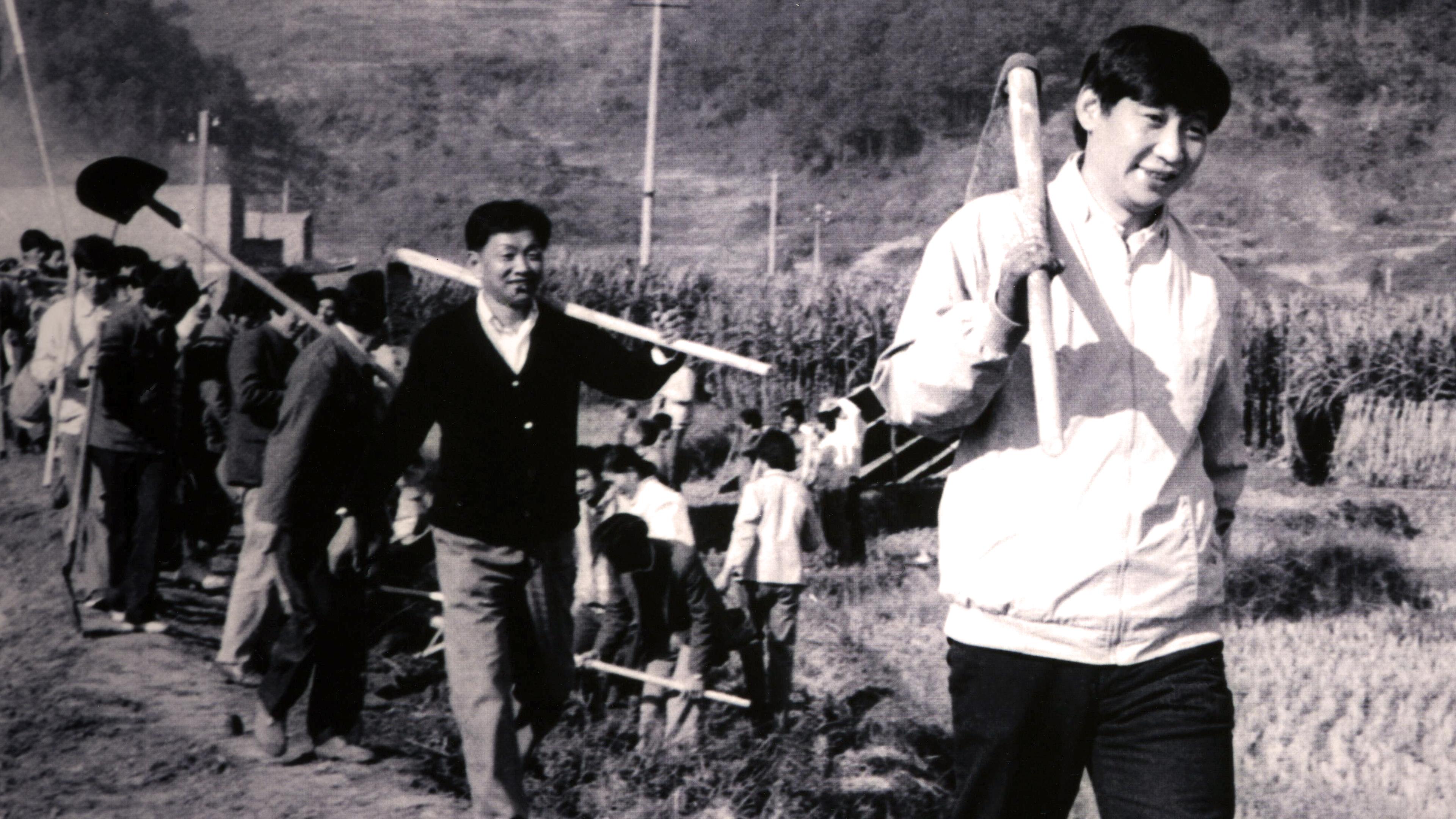 Eine Schwarz-Weiß-Fotoaufnahme von chinesischen Arbeitern, die Schaufeln tragen und in einer Reihe über ein Feld laufen.