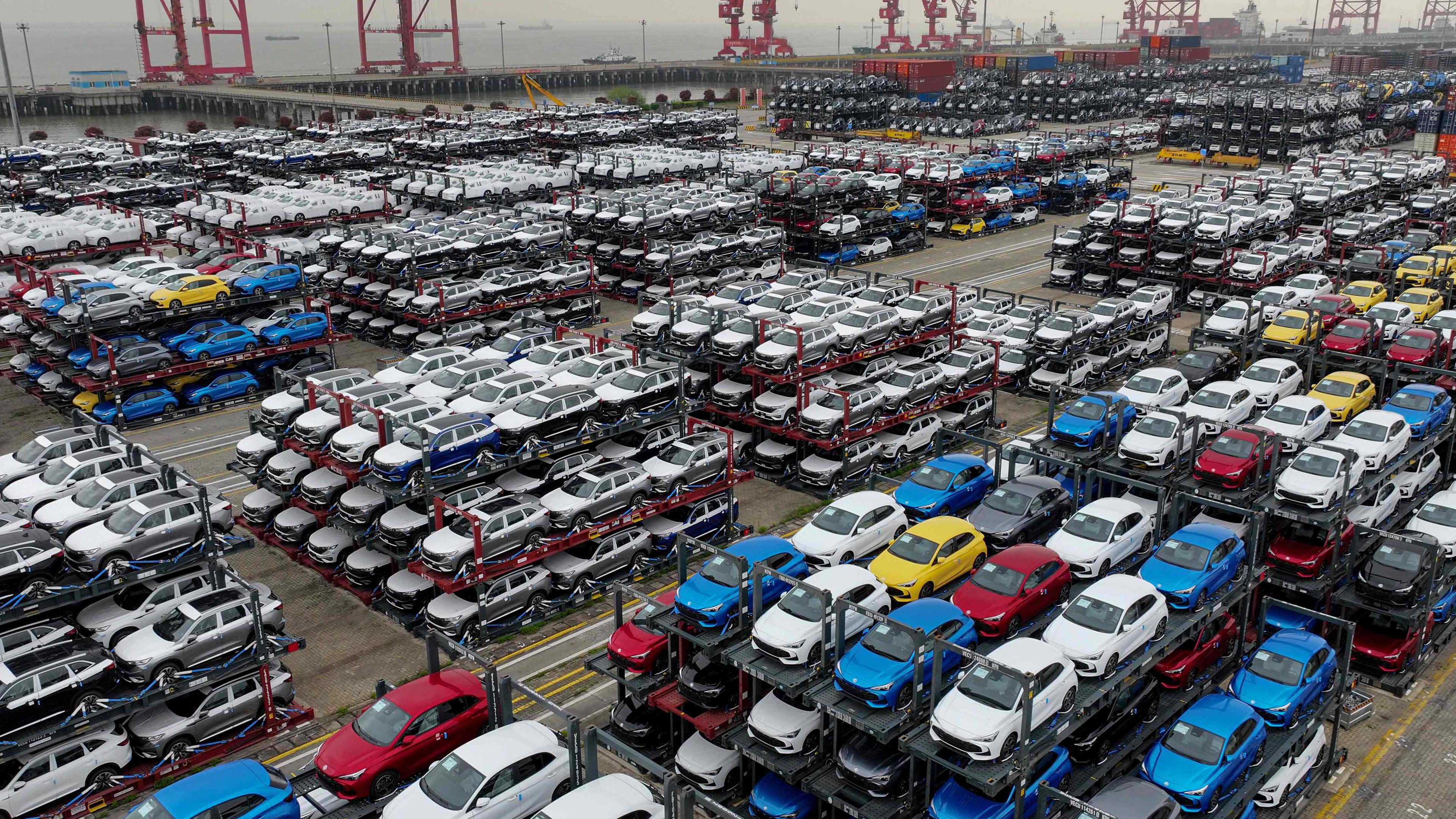 Eine Luftaufnahme von einer Halde mit Elektroautos im Hafen von Suzhou. Im Vordergrund sind zahllose, gestapelte Autos zu sehen, im Hintergrund Wasser und die Kräne eines Hafens.