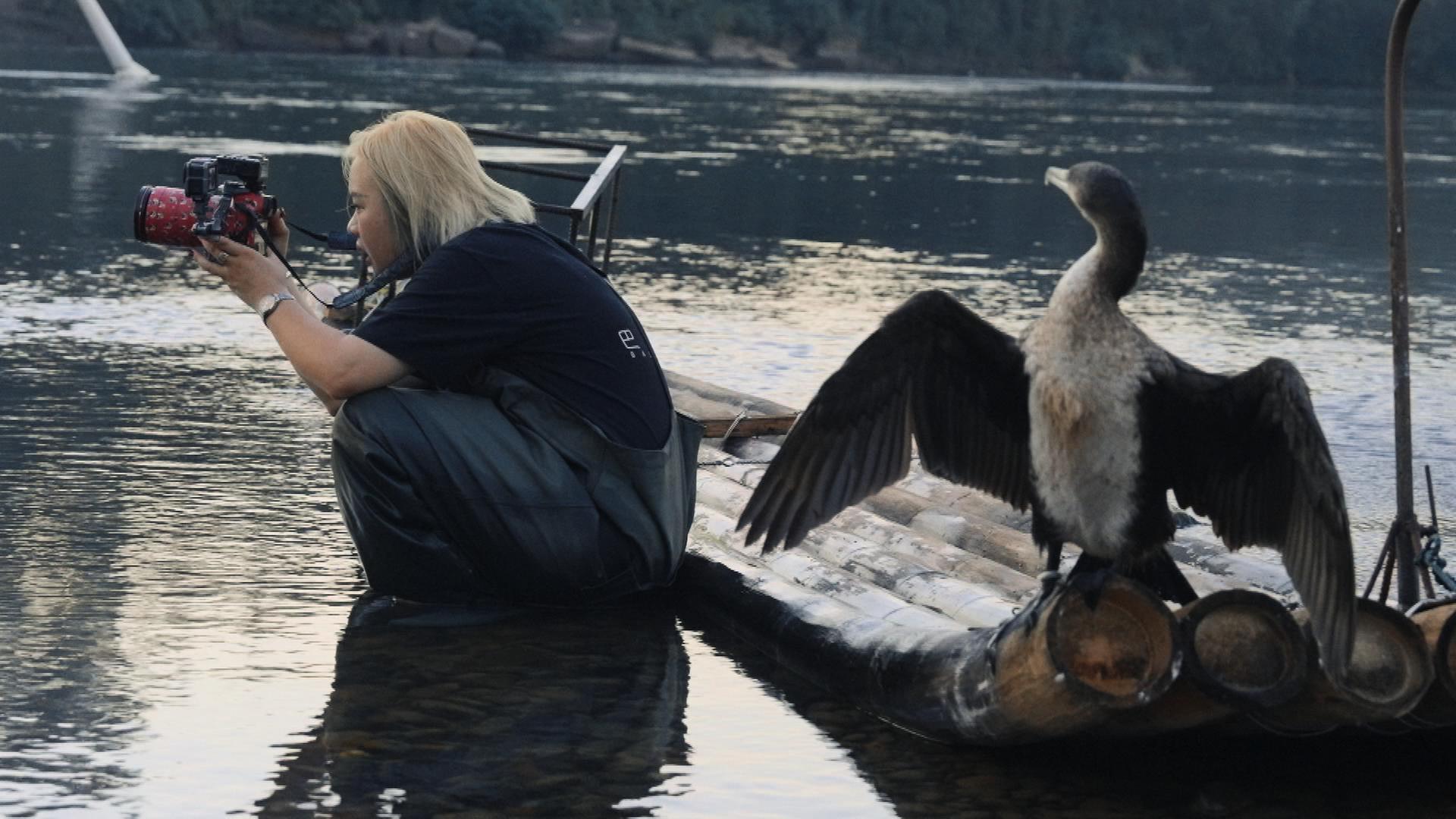 Eine junge Chinesin kniet sich hin, um ein Foto zu schießen. Im Vordergrund spreizt ein Vogel seine Flügel.
