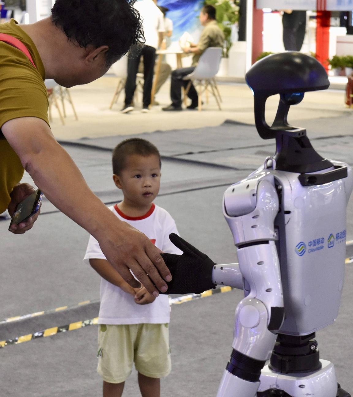 Das Bild ist auf der Global Industrial Conference in Shenyang enstanden. Darauf zu sehen ist ein kleiner Junge, der einen Roboter erstaunt ansieht. Der erwachsene Mann neben ihm gibt dem Roboter die Hand.