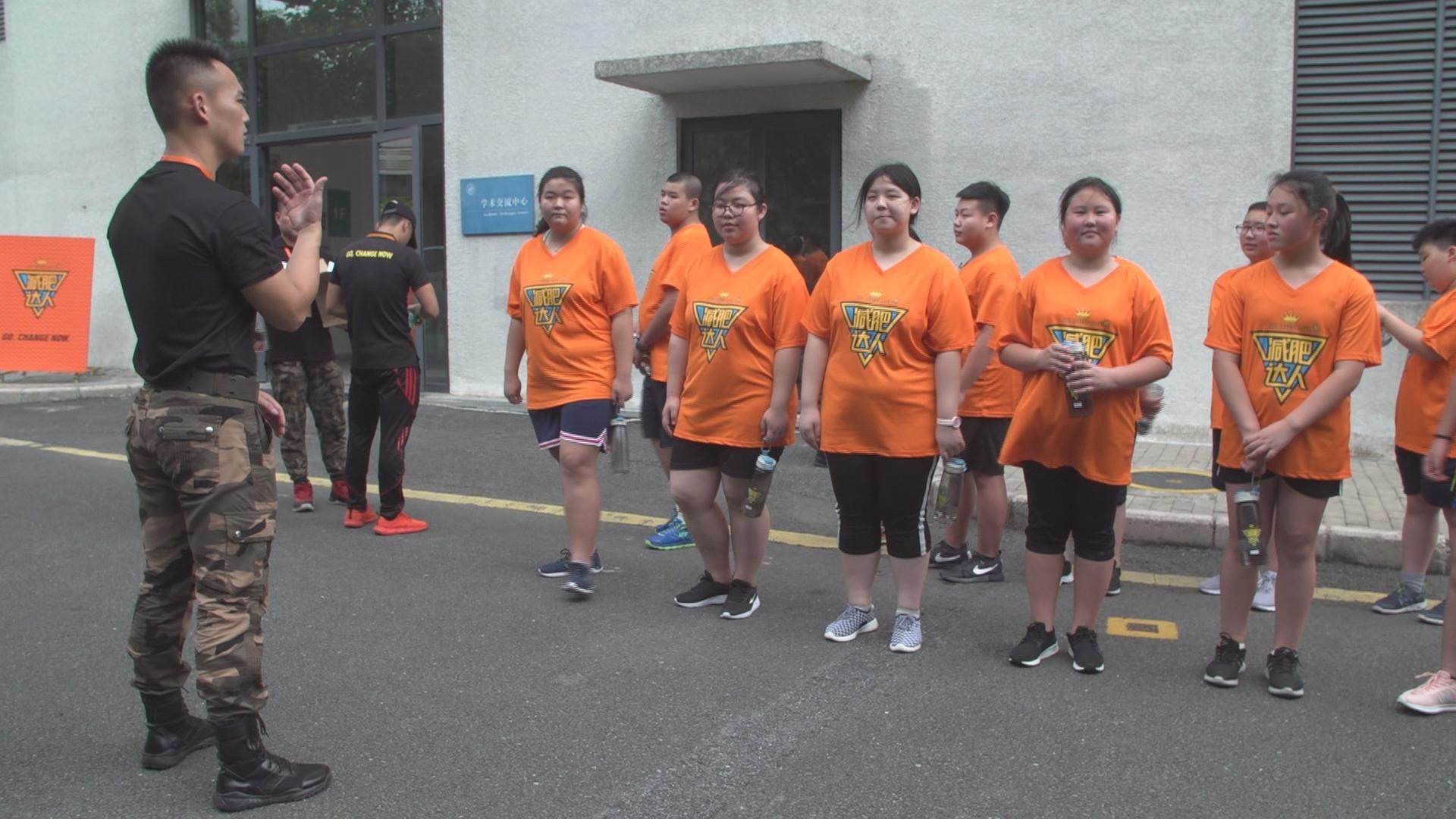 "Chinas dicke Kinder – Die Schattenseite des Wirtschaftsbooms": Eine Gruppe von jugendlichen Chinesen steht in orangefarbenen T-Shirts vor einem Trainer in militärischer Kleidung.