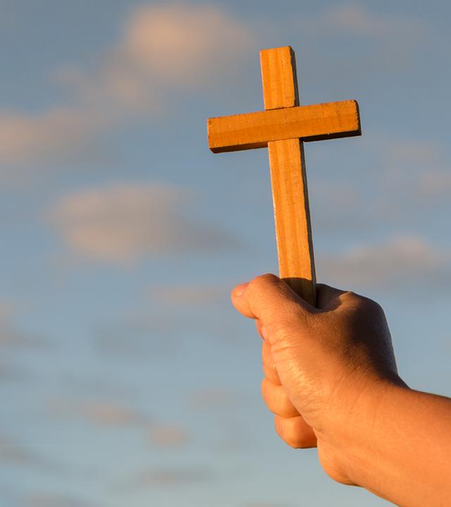 Eine Hand, die ein Holzkreuz vor freiem Himmel hält.
