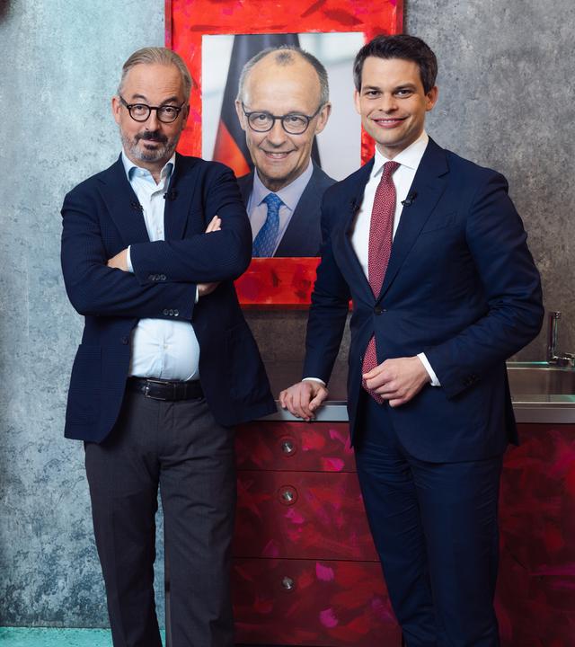 Das Bild zeigt den CDU-Politiker Christoph Ploß und Jan Fleischhauer in einer stilisierten Raumkulisse mit grauer Betonwand. Beide stehen nebeneinander vor einer kleinen Küchenzeile und einem auffällig rot bemalten Kühlschrank. Hinter ihnen hängt ein großformatiges Porträt von Friedrich Merz in einem rot gestalteten Rahmen.
