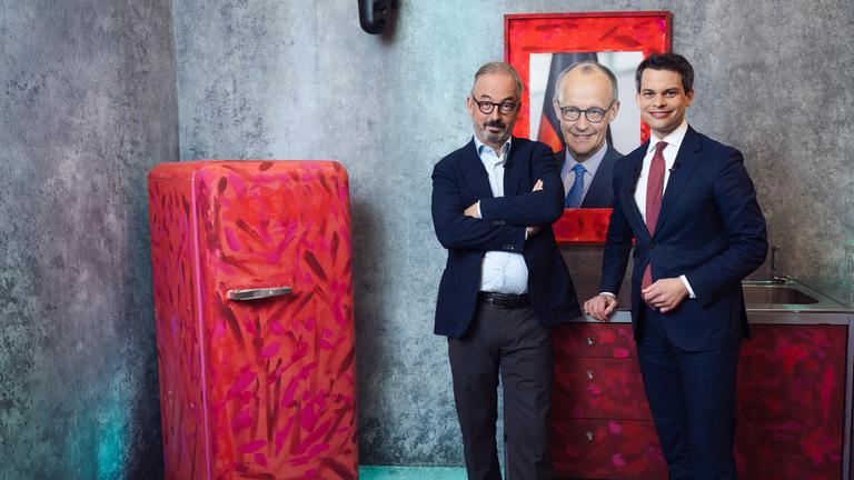 Das Bild zeigt den CDU-Politiker Christoph Ploß und Jan Fleischhauer in einer stilisierten Raumkulisse mit grauer Betonwand. Beide stehen nebeneinander vor einer kleinen Küchenzeile und einem auffällig rot bemalten Kühlschrank. Hinter ihnen hängt ein großformatiges Porträt von Friedrich Merz in einem rot gestalteten Rahmen.