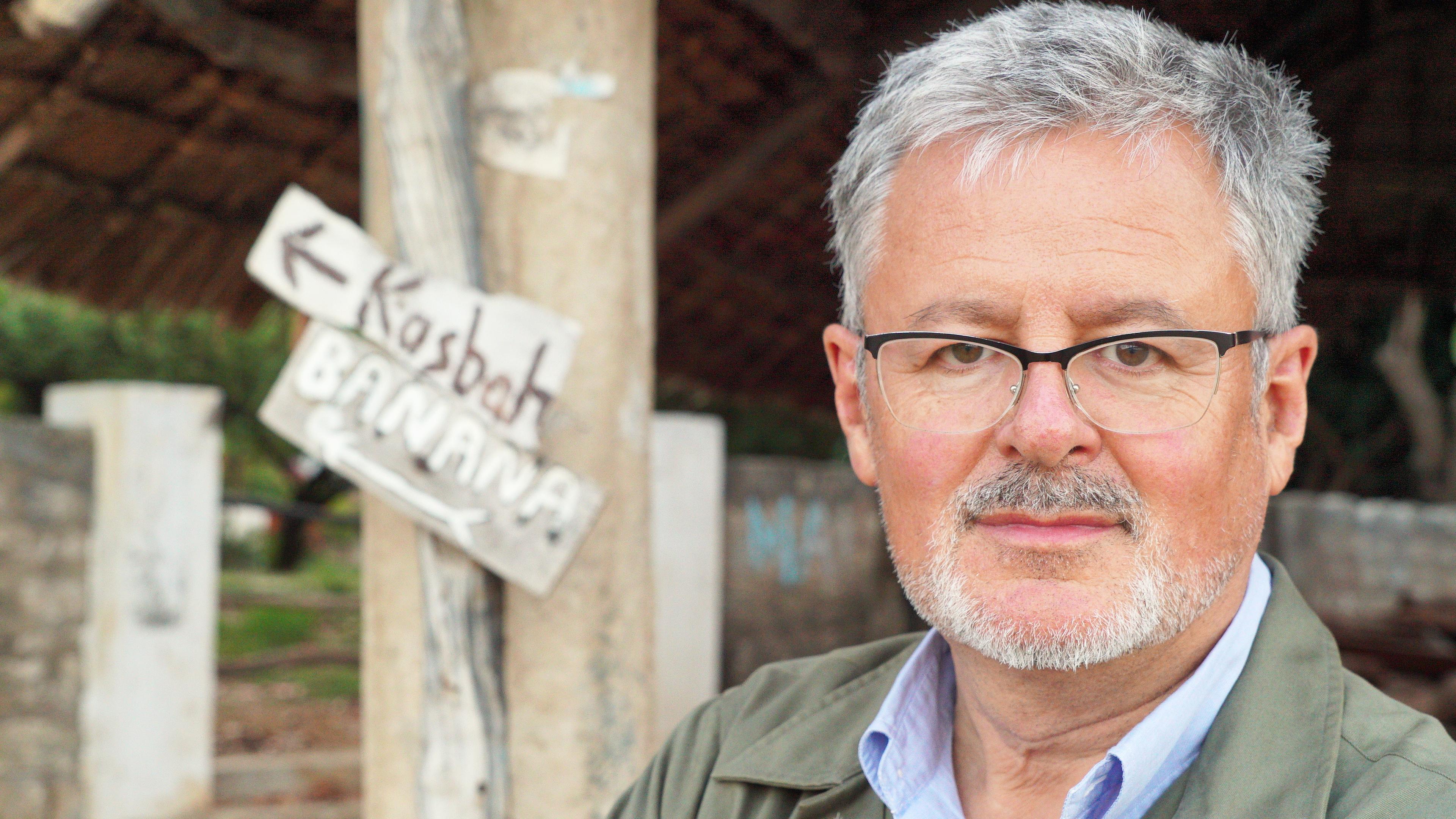 Moderator Christopher Clark im blauen Hemd vor einer afrikanischen Hütte.