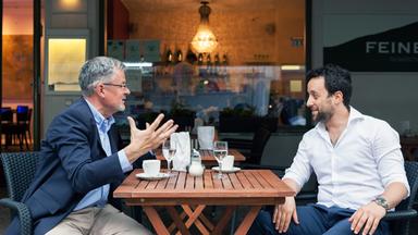Christopher Clark und Yorai Feinberg sitzen sich an einem Café-Tisch im Freien gegenüber und diskutieren.