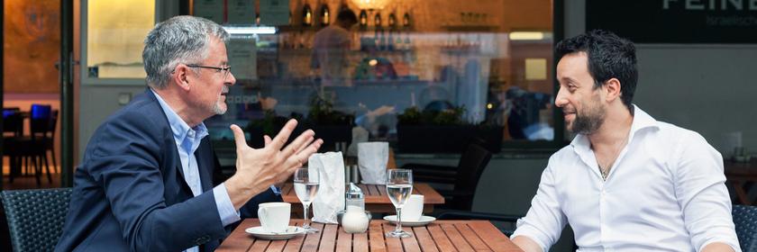 Christopher Clark und Yorai Feinberg sitzen sich an einem Café-Tisch im Freien gegenüber und diskutieren.