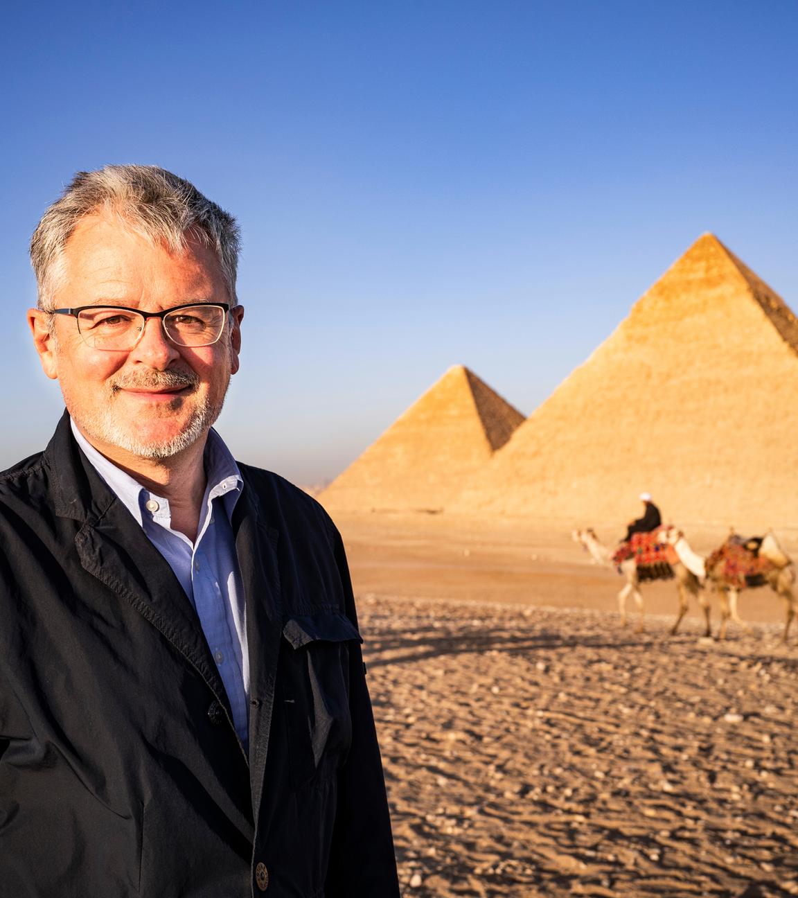 Außenaufnahme von den Pyramiden von Gizeh. Moderator Christopher Clark lächelt in die Kamera. Im Hintergrund ein Mann mit drei Dromedaren.
