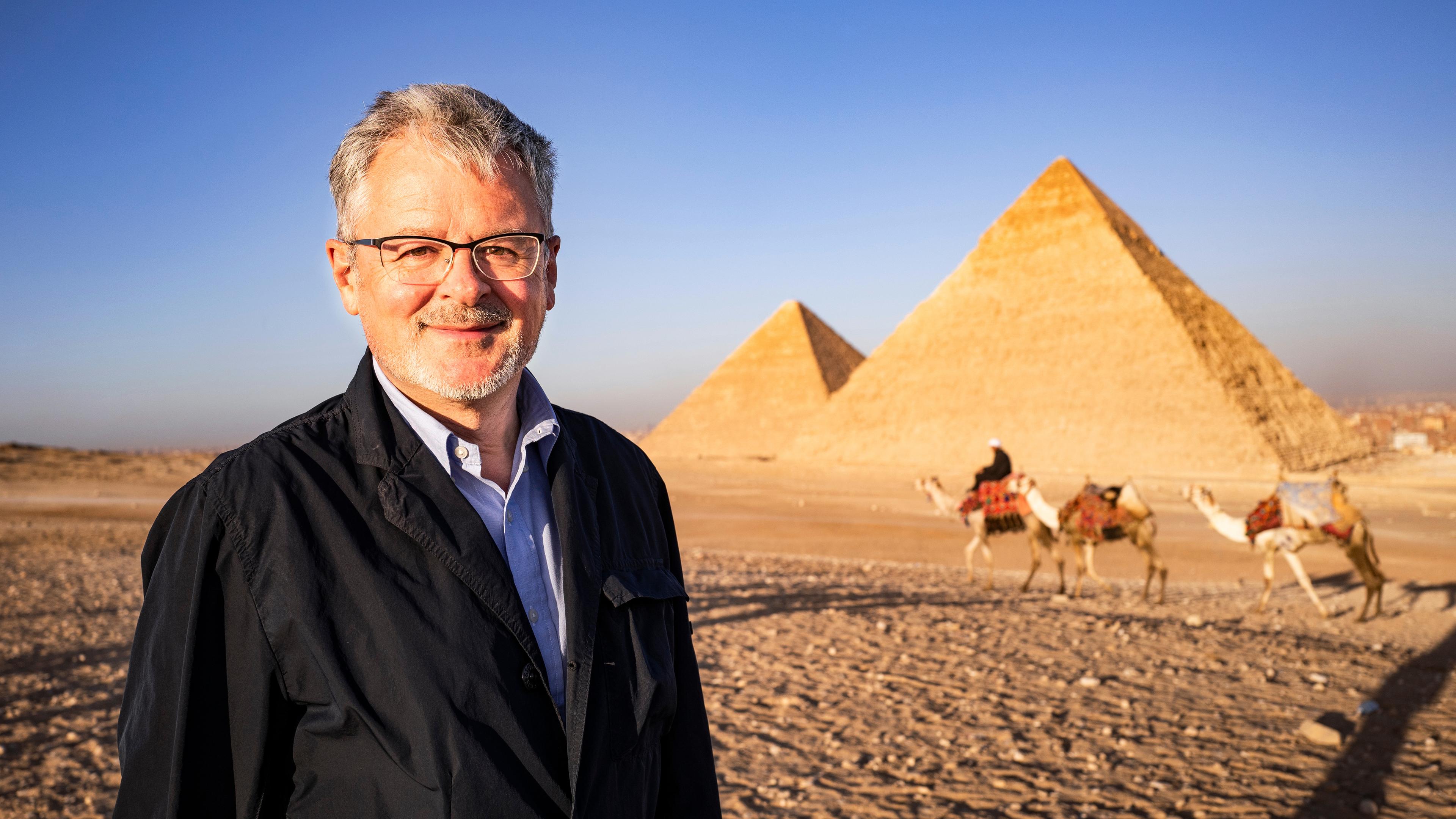Außenaufnahme von den Pyramiden von Gizeh. Moderator Christopher Clark lächelt in die Kamera. Im Hintergrund ein Mann mit drei Dromedaren.