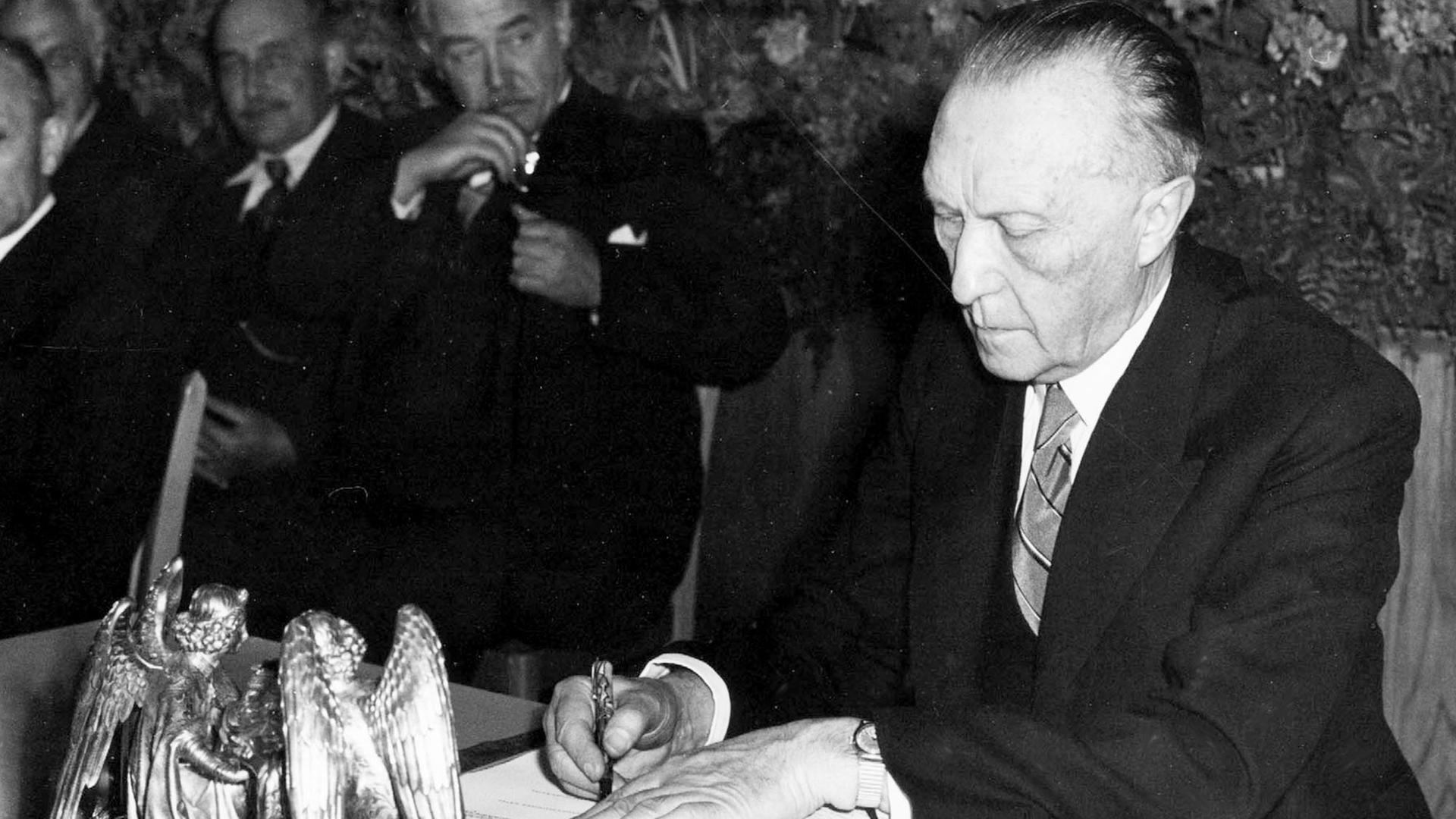 "ZDFzeit: Wir Deutschen und die Demokratie: Schwarzweiß-Foto: Konrad Adenauer bei der Unterzeichnung des Grundgesetzes. Neben ihm auf dem Tisch ein Tintenfass in Engelform.