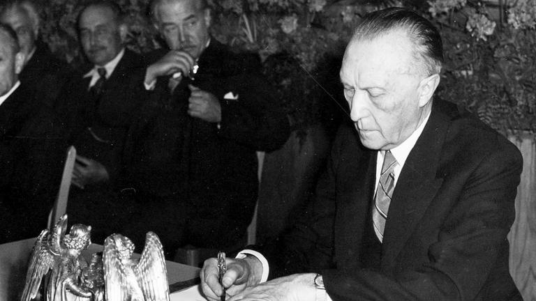 "ZDFzeit: Wir Deutschen und die Demokratie: Schwarzweiß-Foto: Konrad Adenauer bei der Unterzeichnung des Grundgesetzes. Neben ihm auf dem Tisch ein Tintenfass in Engelform.