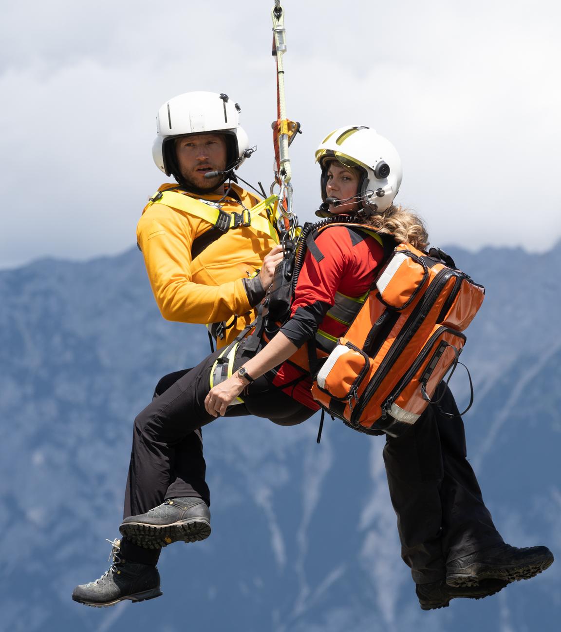 "Die Bergretter: Was wirklich zählt": Die Bergretter Katharina (Luise Bähr) und Markus (Sebastian Ströbel) hängen in ihrer Ausrüstung an einem Seil unter dem Rettungshubschrauber und schweben in der Luft. Im Hintergrund eine Berglandschaft.