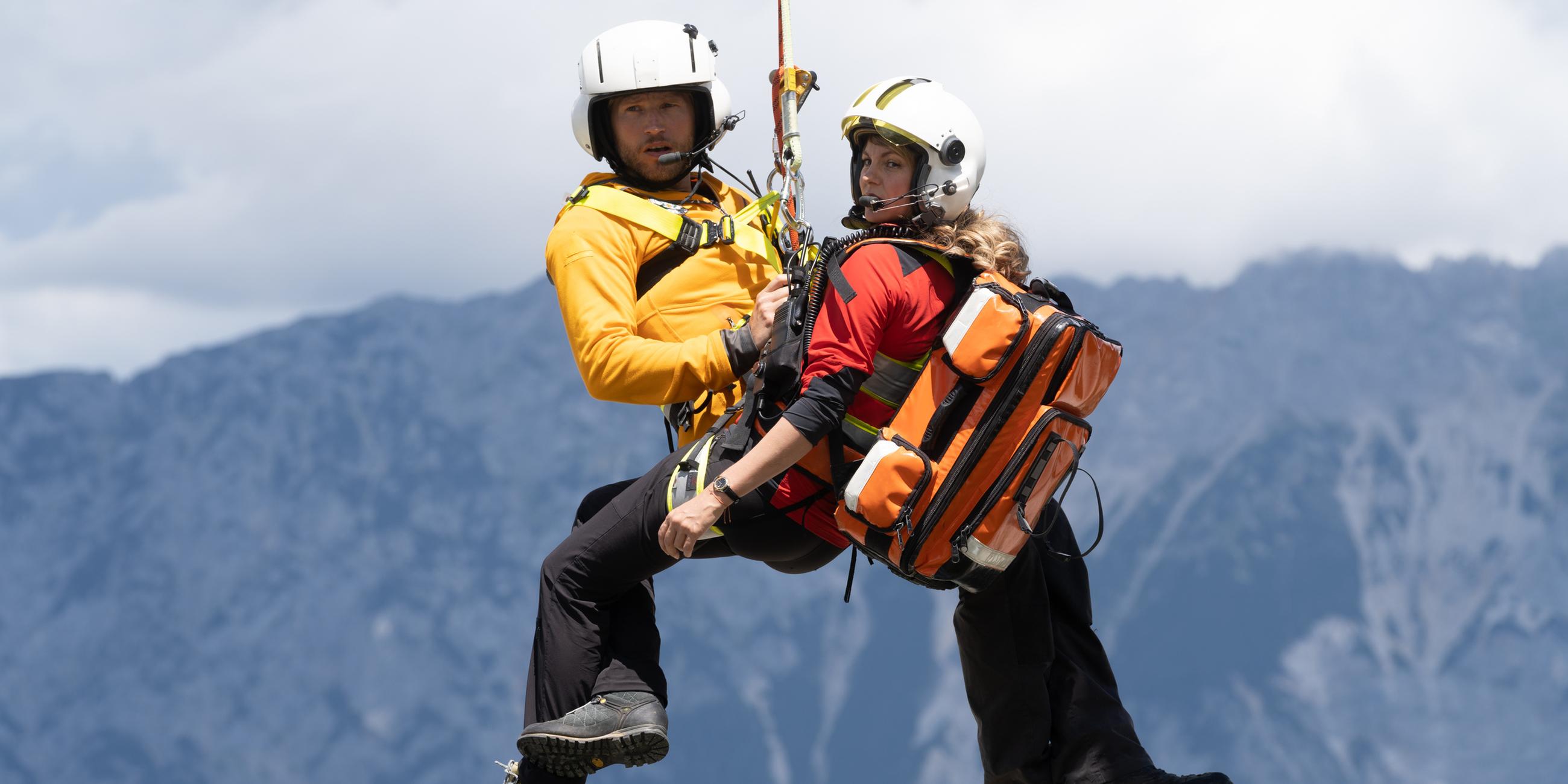 "Die Bergretter: Was wirklich zählt": Die Bergretter Katharina (Luise Bähr) und Markus (Sebastian Ströbel) hängen in ihrer Ausrüstung an einem Seil unter dem Rettungshubschrauber und schweben in der Luft. Im Hintergrund eine Berglandschaft.