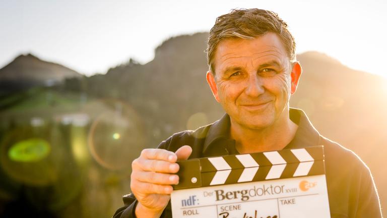 "Backstage - Der Bergdoktor: Am Set mit Hans Sigl": Schauspieler Hans Sigl steht bei Dreharbeiten in einer Berglandschaft. Er hält eine Filmklappe in der Hand und schaut lächelnd in die Kamera.