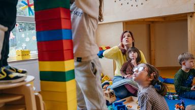 Eine Erzieherin sitzt mit ihren Kindergartenkindern am Boden, zwei Kinder stehen im Vordergrund und bauen einen Turm.