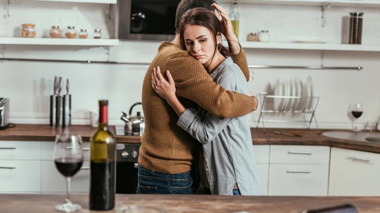 Ein Paar sorgt liegt sich in den Armen. Sie stehen in einer Küche und auf einer Anrichte steht ein Weinglas und eine Weinflasche.