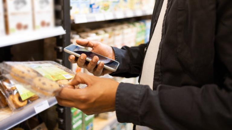 Ein Mann hält eine Packung mit Keksen in der einen Hand, in der anderen ein Smartphone.