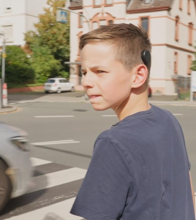 Ein Junge, der ein Cochlea‑Implantat trägt, und gerade die Straße überqueren möchte.