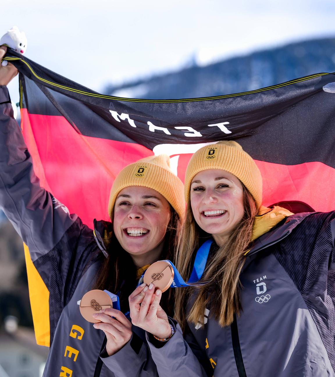 Die deutschen Skilangläuferinnen Coletta Rydzek und Laura Gimmler präsentieren jubelnd ihre Bronzemedaillen und halten zusammen die Flagge Deutschlands hoch