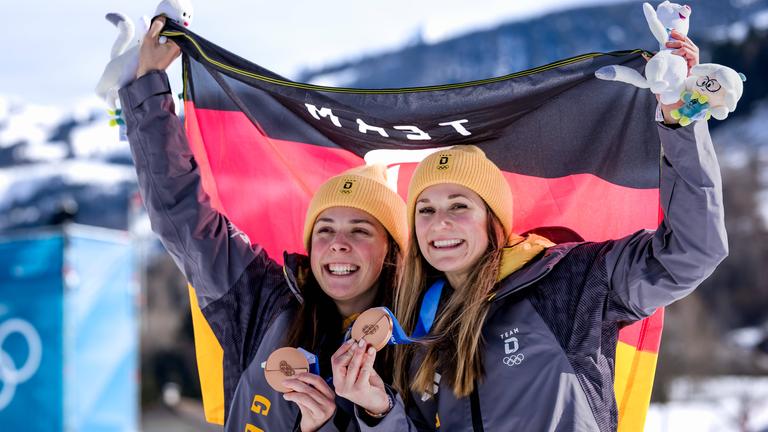 Die deutschen Skilangläuferinnen Coletta Rydzek und Laura Gimmler präsentieren jubelnd ihre Bronzemedaillen und halten zusammen die Flagge Deutschlands hoch