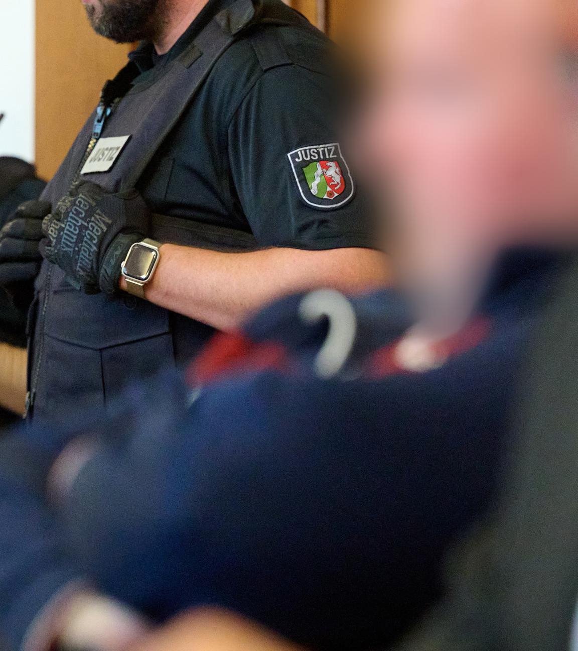 Einer der Angeklagten sitzt neben seinem Anwalt, während im Hintergrund ein Polizist steht.