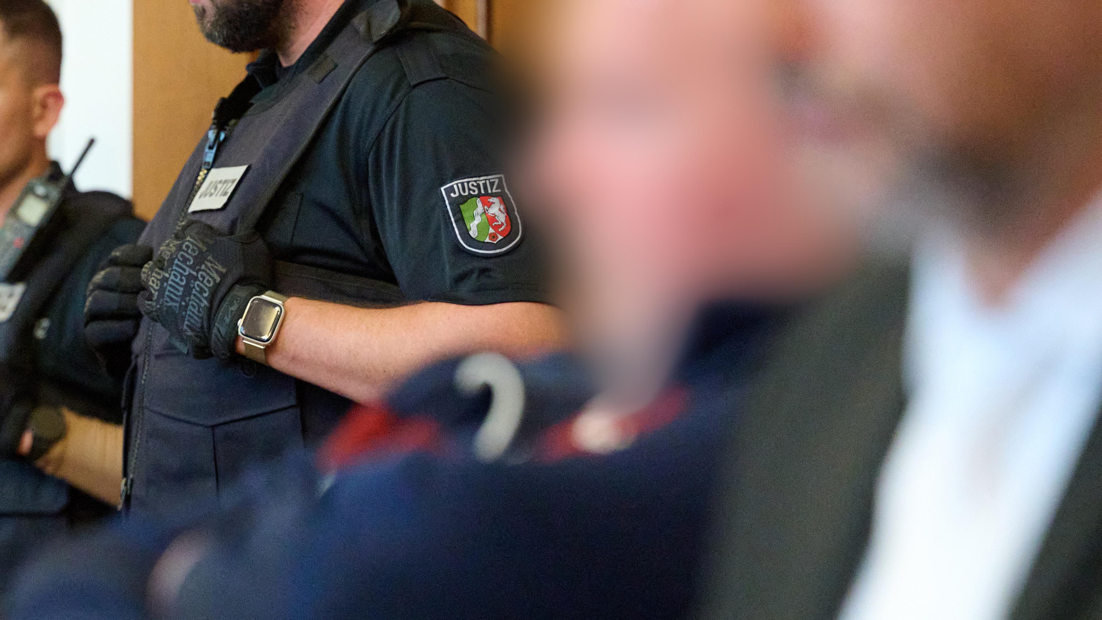 Einer der Angeklagten sitzt neben seinem Anwalt, während im Hintergrund ein Polizist steht.