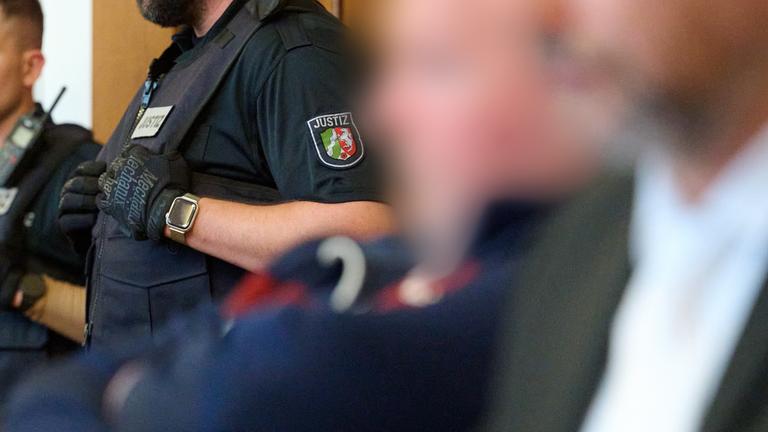 Einer der Angeklagten sitzt neben seinem Anwalt, während im Hintergrund ein Polizist steht.