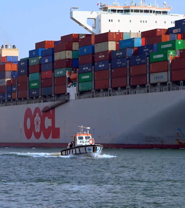 Ein großer Frachter mit bunten Containern, davor ein kleines Lotsenboot.