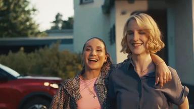 Cosmia Henman und Klara Lange stehen vor einem Gebäude. Cosima hat den Arm um die Schultern von Klara gelegt. Im Hintergrund ist ein rotes Fahrzeug zu sehen.