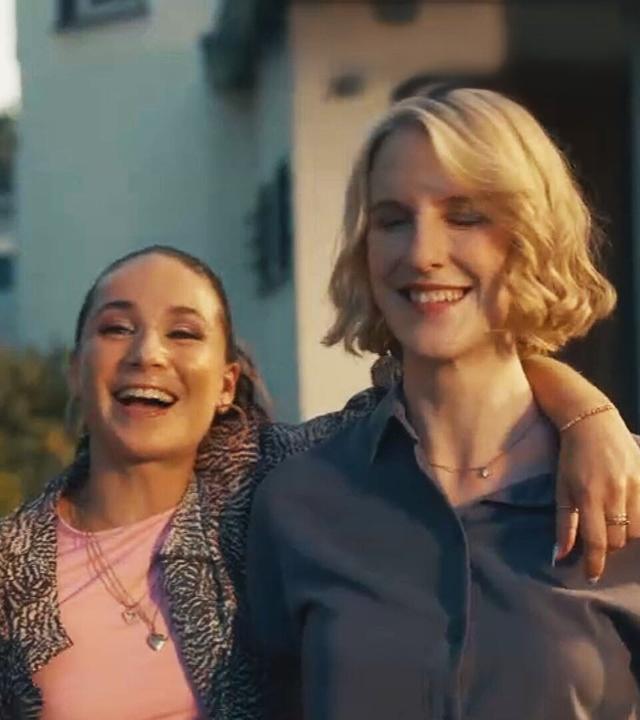 Cosmia Henman und Klara Lange stehen vor einem Gebäude. Cosima hat den Arm um die Schultern von Klara gelegt. Im Hintergrund ist ein rotes Fahrzeug zu sehen.