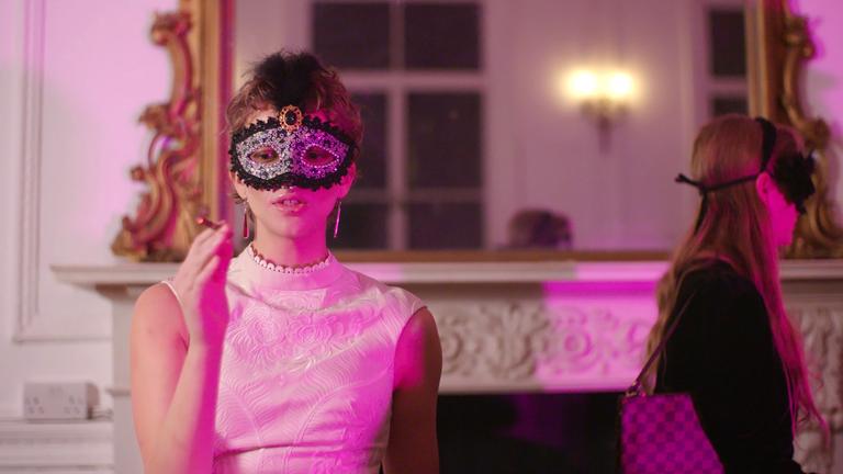 Eine kurzhaarige Frau in weißem Abendkleid und mit einer auffällig verzierten Augenmaske sitzt vor einem offenen Kamin. In der einen Hand hält sie eine Zigarette.